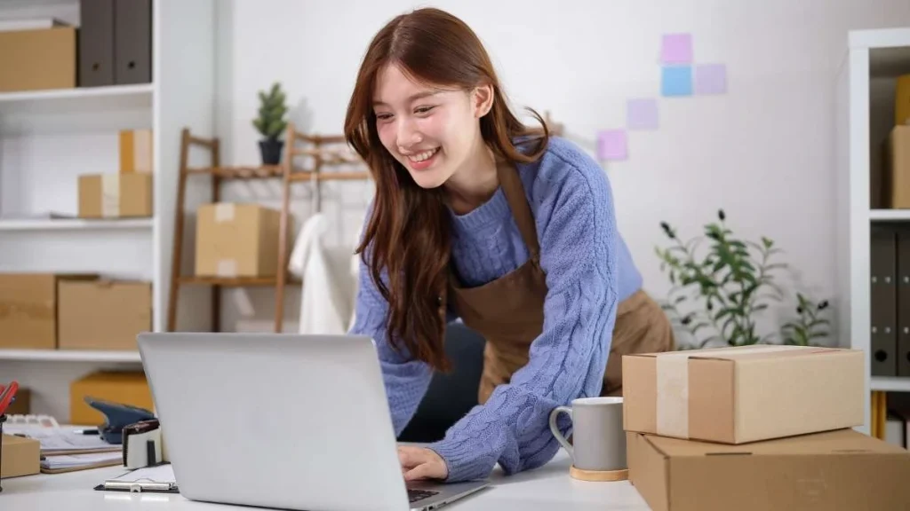 1 - Gambar seorang wanita sedang membuka laptop dengan paket di sisinya