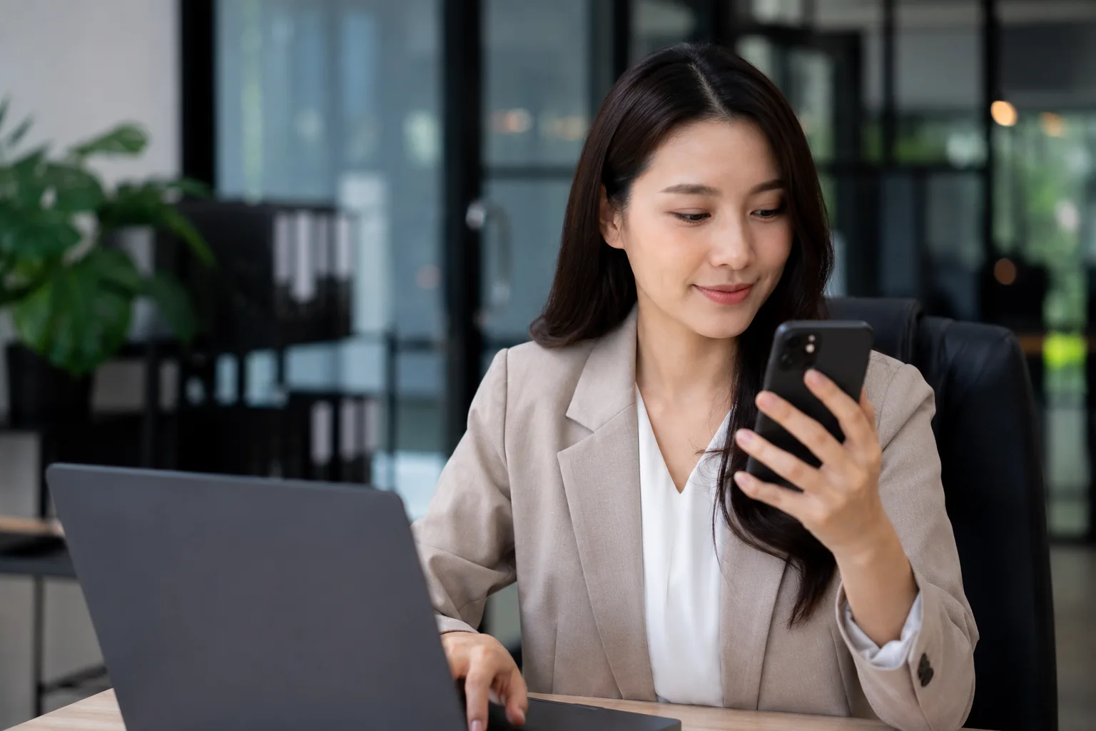 perempuan bisnis mengenakan blazer memeriksa ponsel di samping laptop